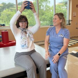 Patient lifts small exercise ball during a cancer rehab physical therapy session while the therapist watches and guides. 